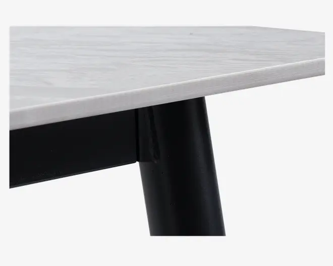 Nærbillede af et bordhjørne, der viser en glat, lysefarvet bordplade med diskret træstruktur. Bordpladen har en lige kant og er understøttet af et robust, cylindrisk sort metalben, der er fastgjort underneden.