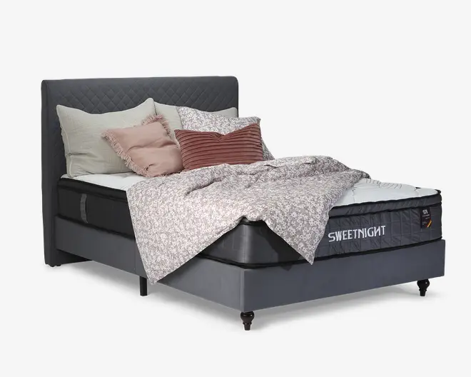 Et moderne sengesæt med et quiltet, mørkegråt polstret hovedgærde og matchende base. Madrassen har en mellemtykkelse med sort og hvid farvekombination samt synligt 'SWEETNIGHT'-mærke på siden. Sengen er pyntet med flere dekorative puder i creme, lyserød og stribet rød, samt et lyst, blomster-mønstret dynebetræk, der ligger afslappet hen over madrassen. Sengen står på korte, skrå ben.