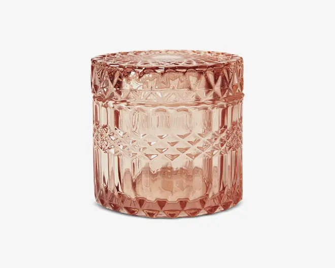 Rund beholder lavet af lyserødt glas med tilhørende låg. Overfladen har et indviklet, hævet geometrisk mønster med diamant- og stjerneformer, hvilket giver et vintage og dekorativt udtryk. Glasset er tykt og reflekterende, hvilket tilføjer et luksuriøst præg.