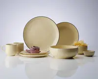 Et sæt matchende keramikservice i cremefarve med blank overflade, bestående af to store middagstallerkener, en mindre frokosttallerken, en suppeskål, en lille skål, to krus og en sidetallerken. Delene har en diskret, tynd mørk kant langs kanten, hvilket giver et elegant udtryk. Sættet er præsenteret på en reflekterende overflade, og en tallerken er pyntet med en sammenfoldet, lilla serviet. Designet er minimalistisk og moderne.