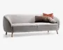 En moderne sofa med lyst gråt, tekstureret stofbetræk og blødt buede armlæn. Sofaen står på fire lyse træben, hvilket giver den et skandinavisk udtryk. Den er pyntet med tre pyntepuder: en rektangulær brun, fluffy pude, en rund, plisseret lysegrå pude og en kvadratisk, rustfarvet pude, som tilføjer både komfort og stil.