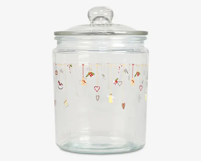 Klar glasbeholder med rundt låg og knop som håndtag. Beholderen har et festligt design øverst med små illustrationer af julemotiver som slikstokke, hjerter, gaver, engle og stjerner, der hænger ned fra gyldne linjer. Glasset er cylindrisk og har en glat, gennemsigtig overflade.