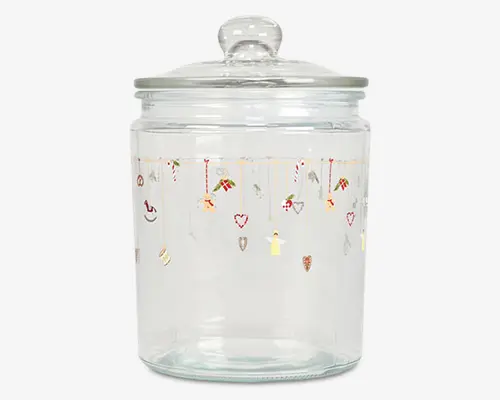 Klar glasbeholder med rundt låg og knop som håndtag. Beholderen har et festligt design øverst med små illustrationer af julemotiver som slikstokke, hjerter, gaver, engle og stjerner, der hænger ned fra gyldne linjer. Glasset er cylindrisk og har en glat, gennemsigtig overflade.