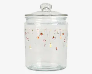 Klar glasbeholder med rundt låg og knop som håndtag. Beholderen har et festligt design øverst med små illustrationer af julemotiver som slikstokke, hjerter, gaver, engle og stjerner, der hænger ned fra gyldne linjer. Glasset er cylindrisk og har en glat, gennemsigtig overflade.
