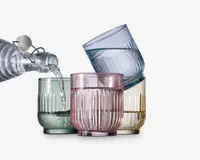 Sæt med fire lave drikkeglas, hver med rillet tekstur og i forskellige gennemsigtige farver: grøn, pink, blå og gul. Glassene har en tyk bund og lige sider. Vand hældes fra en klar glasflaske med patentlåg ned i det grønne glas.