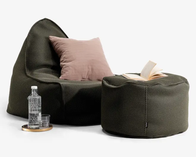 Et moderne loungemiljø med en mørke olivengrøn, tekstureret sækkestol og en matchende puf. Sækkestolen har en blød, struktureret form og er pyntet med en stor, lyse lilla pude. På puffen ligger en åben bog. Ved siden af stolen står et rundt træfad med en klar glasflaske og to glas, hvilket giver et praktisk og stilfuldt udtryk.