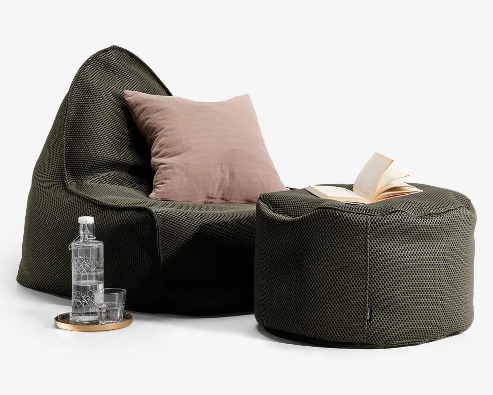 Et moderne loungemiljø med en mørke olivengrøn, tekstureret sækkestol og en matchende puf. Sækkestolen har en blød, struktureret form og er pyntet med en stor, lyse lilla pude. På puffen ligger en åben bog. Ved siden af stolen står et rundt træfad med en klar glasflaske og to glas, hvilket giver et praktisk og stilfuldt udtryk.