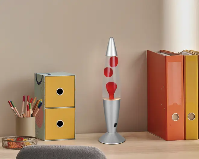 En sølvfarvet og gennemsigtig lampe med klassisk lavalampe-design, der indeholder røde voksbobler i klar væske. Lampen har en konisk top og en bred, rund fod. Den står på et lyst træskrivebord ved siden af et sæt gule og blå opbevaringsskuffer, en beige kop med farveblyanter og flere orange og gule ringbind. Lampens moderne metalfinish og de levende røde voksbobler giver et retro, men samtidig moderne udtryk.