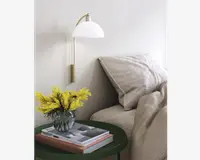 Et moderne soveværelsesmiljø med en seng, der er redt med beige, tekstureret sengetøj og store puder. Ved siden af sengen står et rundt, mørkegrønt natbord med en klar glasvase med lysegule blomster og en stak hardback-bøger. Over bordet hænger en vægmonteret lampe med messingarm og hvid, kuppelformet skærm, der giver blødt lys. Udtrykket er rent, hyggeligt og moderne.