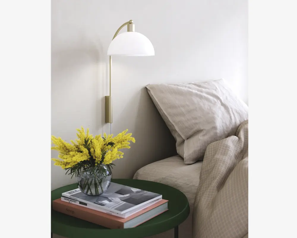 Et moderne soveværelsesmiljø med en seng, der er redt med beige, tekstureret sengetøj og store puder. Ved siden af sengen står et rundt, mørkegrønt natbord med en klar glasvase med lysegule blomster og en stak hardback-bøger. Over bordet hænger en vægmonteret lampe med messingarm og hvid, kuppelformet skærm, der giver blødt lys. Udtrykket er rent, hyggeligt og moderne.