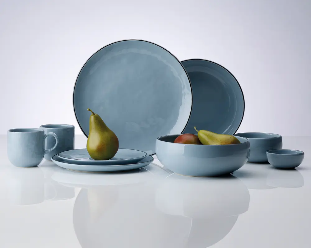 Et sæt lyseblåt keramikstel med blank overflade, bestående af to store middagstallerkener, to mindre tallerkener, to krus, en stor serveringsskål, en mellemstor skål og en lille skål. Delene har en let uregelmæssig form og en tynd, mørkere kant. Sættet er præsenteret på en reflekterende hvid overflade, og nogle af delene indeholder frugt for at vise størrelsen.