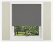 Et vindue med en mørkegrå rullegardin, der er delvist rullet ned og dækker den øverste halvdel af vinduet. Gardinet har et glat, ensfarvet stof uden synlige mønstre eller teksturer. Vinduesrammen er hvid, og væggen omkring er lysebeige. Den nederste del af vinduet giver udsyn til grønne træer og græs udenfor.