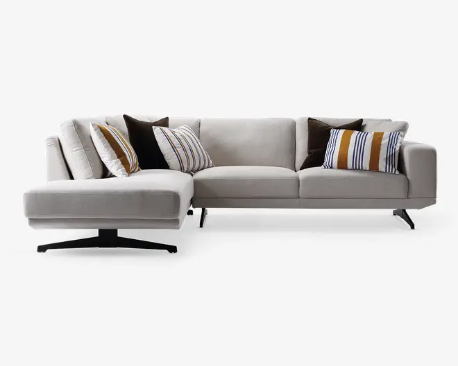 Moderne L-formet sofa betrukket med lysebeige stof, med brede armlæn og slanke sorte metalben. Sofaen har flere dekorative puder: nogle med brune og sennepsgule striber, andre i ensfarvet mørkebrun, samt et par med flerfarvede striber, hvilket giver et stilfuldt og hyggeligt udtryk.