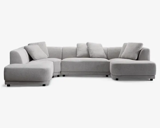 En stor, moderne hjørnesofa betrukket med lysegråt stof. Sofaen har et rummeligt L-formet design med brede, dybe sæder og lavt ryglæn. Den leveres med fem matchende, bløde ryghynder for ekstra komfort. Armlænene er brede og let afrundede, og stilen er minimalistisk med rene linjer og bløde kanter.