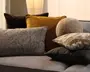 En samling dekorative puder i blandede ტექsturer og farver, herunder mørkebrun velour, sennepsgul, grå vævet stof, en rund beige plys-pude og mønstrede pyntepuder på en lysegrå sofa.