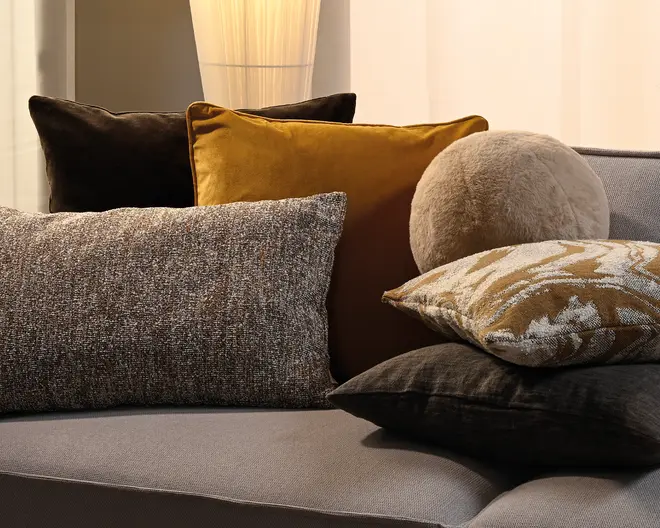 En samling dekorative puder i blandede ტექsturer og farver, herunder mørkebrun velour, sennepsgul, grå vævet stof, en rund beige plys-pude og mønstrede pyntepuder på en lysegrå sofa.