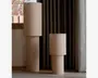 To minimalistiske beige cylindriske elementer med glatte, matte overflader, én høj og én lavere, hver sammensat af tragt- eller trommelignende sektioner i et blødt oplyst rum ved et vindue og et gardin.