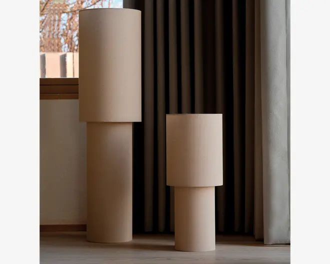 To minimalistiske beige cylindriske elementer med glatte, matte overflader, én høj og én lavere, hver sammensat af tragt- eller trommelignende sektioner i et blødt oplyst rum ved et vindue og et gardin.