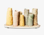 Et dekorativt keramikstykke bestående af otte opretstående, cylindriske rør i forskellige højder og diametre, alle fastgjort til en oval base. Hvert rør er unikt glaseret i pastelfarver som gul, lyserød, blå, grøn og creme, og har indgraverede blomster- og snirklemønstre. Rørene har let ujævne, organiske former og skrå åbninger, hvilket giver et håndlavet, kunstnerisk udtryk. Basen er plettet og matcher farverne på rørene.