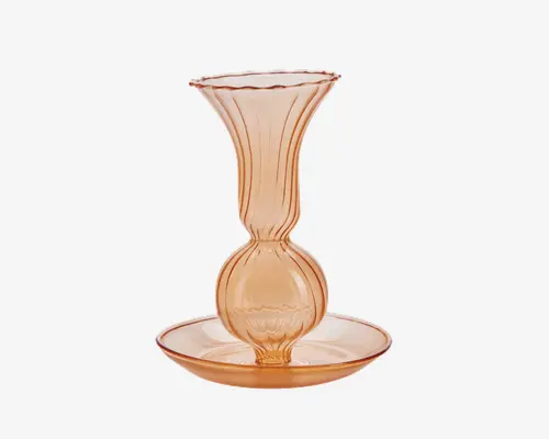 Gennemsigtigt glas i ferskenfarve med en bred, bølget kant og lodrette rillede detaljer. Kroppen har en rund, kugleformet midte og står på en bred, lav skålformet fod. Glasset har en glat, blank overflade og et elegant, vintage-inspireret design.