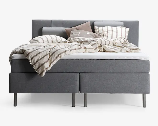 En moderne seng med gråt polstret stel og sengegavl, boksmadras og metalben. Sengen er redt med et beige og hvidt stribet dynebetræk og matchende puder samt ekstra pyntepuder i neutrale farver. Sengetøjet ser blødt og indbydende ud med et let rodet, afslappet udtryk.
