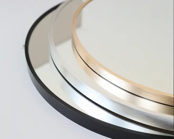 Nærbillede af tre runde genstande med metalrammer i forskellige finish: mat sort, blank sølv og børstet guld. Rammerne er slanke og moderne med en glat, reflekterende overflade. Genstandene er delvist stablet oven på hinanden, hvilket fremhæver kontrasten mellem de forskellige rammefarver og finish.