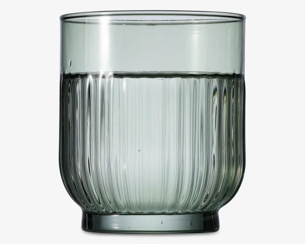 Kort drikkeglas med let afrundet bund og lodret rillet struktur på den nederste halvdel. Glasset er transparent med et diskret gråt skær, og kanten er glat og jævn. Designet giver et moderne og elegant udtryk, velegnet til servering af vand eller andre drikkevarer.