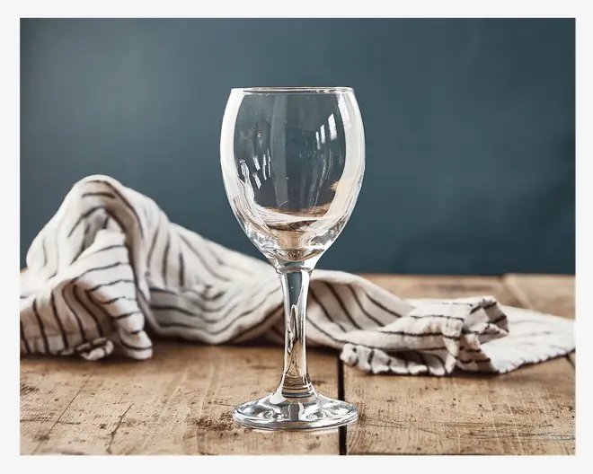 Et klart glas med stilk, rundet skål og let udadbuet kant, velegnet til servering af vin. Glasset står på en rustik træoverflade med en stribet stofserviet i baggrunden, hvilket fremhæver dets gennemsigtighed og klassiske design.