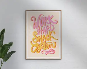 Et indrammet print med teksten 'Work Hard Snack Often' i fed, legende håndskrevet skrift. Teksten er skrevet i nuancer af pink, gul og orange på en lys beige baggrund, med dekorative krøller og en diskret hvid detalje. Rammen er enkel og i lyst træ, og printet er udstillet mod en tekstureret hvid væg med en grøn plante delvist synlig til venstre.