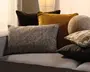 En lagdelt opstilling af dekorative puder i mørkebrun, sennepsgul, meleret grå, beige og sort med en blanding af velour-, vævede og bløde pelsagtige teksturer på en grå sofa.
