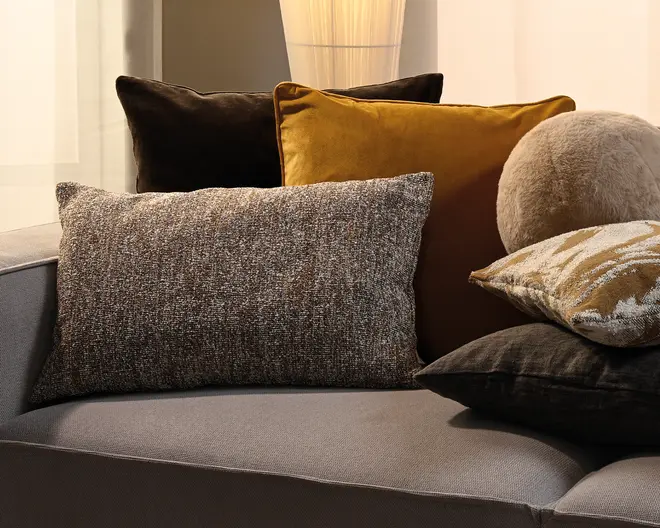 En lagdelt opstilling af dekorative puder i mørkebrun, sennepsgul, meleret grå, beige og sort med en blanding af velour-, vævede og bløde pelsagtige teksturer på en grå sofa.