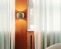 Et moderne vægmonteret lysarmatur med en glat, afrundet rektangulær form og mat finish. Armaturet udsender et varmt, diffust lys både opad og nedad, hvilket skaber en blød atmosfære. Det er installeret på en trævæg ved siden af et vindue med hvide, gennemsigtige gardiner, hvilket bidrager til et rent og moderne interiørdesign.