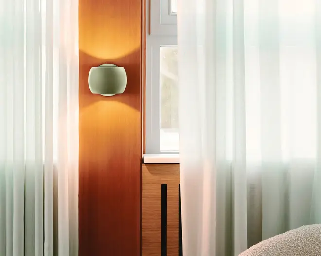 Et moderne vægmonteret lysarmatur med en glat, afrundet rektangulær form og mat finish. Armaturet udsender et varmt, diffust lys både opad og nedad, hvilket skaber en blød atmosfære. Det er installeret på en trævæg ved siden af et vindue med hvide, gennemsigtige gardiner, hvilket bidrager til et rent og moderne interiørdesign.