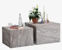 To kubeformede borde med et naturstenslook i grå og beige nuancer, med en glat, mat overflade og synlige åretegninger. Bordene er dekoreret med forskellige pyntegenstande, herunder en potteplante med hvide blomster, glaslysestager, en vandflaske, en bog med briller samt en samling af glaslysestager og vaser. Bordene har et moderne, minimalistisk design med rene linjer og et solidt, robust udtryk.