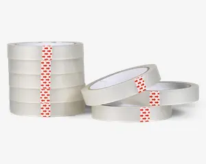 Flere ruller klar, gennemsigtig tape med hvid kerne vises. Hver rulle har en rød strimmel med pilemønster, der holder tapeenden fast. Tapen er blank og gennemsigtig, og rullerne er stablet i to grupper, hvor nogle ligger fladt, og andre står oprejst.