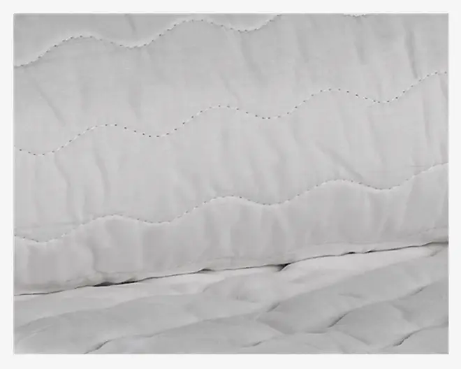 Nærbillede af en hvid quiltet stofoverflade med bølget syningsmønster. Materialet ser blødt og polstret ud og giver en behagelig tekstur. Syningerne er jævnt fordelt og danner et ensartet bølgemønster over stoffet.