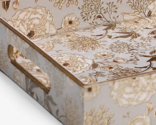 Rektangulær bakke med udskårne håndtag på siderne, dekoreret med et elegant, præget blomsterdesign i guld på en blank hvid baggrund. Blomster og blade er detaljeret udført, hvilket giver overfladen en tekstureret og luksuriøs fornemmelse. Kanterne er afsluttet med en guldfarvet kant, som understreger det sofistikerede udtryk.