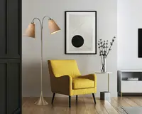 Et moderne stuearrangement med en lys gul lænestol, der har en firkantet silhuet, let buede armlæn og sorte, tilspidsede ben. Stolen har en blød sædepude og en støttende ryg. Ved siden af stolen står en elegant gulvlampe med børstet metalfinish og to justerbare, kegleformede lampeskærme i stof. Lampen har en bred, rund fod. Bag stolen hænger et minimalistisk indrammet kunstværk med geometriske former på en lysegrå væg. Et lille rundt sidebord med hvid bordplade og slanke guldfarvede ben har en høj glasvase med dekorative grene.