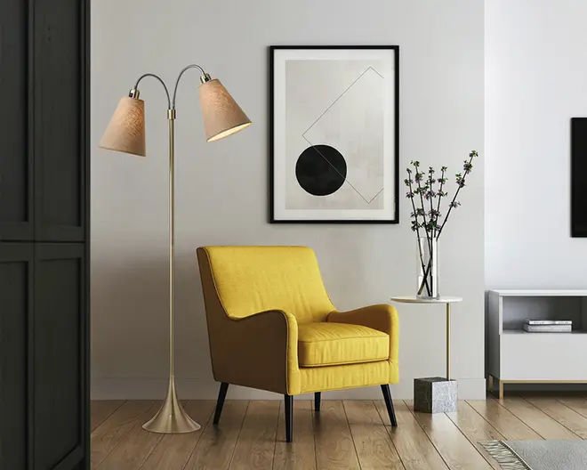 Et moderne stuearrangement med en lys gul lænestol, der har en firkantet silhuet, let buede armlæn og sorte, tilspidsede ben. Stolen har en blød sædepude og en støttende ryg. Ved siden af stolen står en elegant gulvlampe med børstet metalfinish og to justerbare, kegleformede lampeskærme i stof. Lampen har en bred, rund fod. Bag stolen hænger et minimalistisk indrammet kunstværk med geometriske former på en lysegrå væg. Et lille rundt sidebord med hvid bordplade og slanke guldfarvede ben har en høj glasvase med dekorative grene.