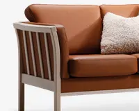 Nærbillede af en sofa med glatte, lysebrune læderhynder og en solid træramme med lys finish. Armlænet og sidepanelet har lodrette trælameller. En stor, blød, cremefarvet pude med tekstur ligger på sædet og giver et hyggeligt præg.