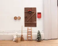 En miniaturetrædør med skråstiver og en lille krans med rød sløjfe er monteret på en hvid væg. Ved siden af døren hænger en lille rød postkasse med teksten 'POST.' Under døren står en lille træstige, en miniaturekost, en kurv, en sæk mærket 'FAIRY' og et lille kunstigt træ med hvide spidser. To små runde træknager er også synlige på væggen. Scenen er opsat som en eventyrlig fe- eller nissedør.
