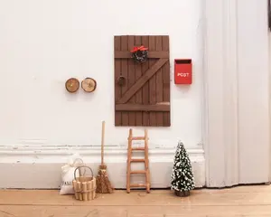 En miniaturetrædør med skråstiver og en lille krans med rød sløjfe er monteret på en hvid væg. Ved siden af døren hænger en lille rød postkasse med teksten 'POST.' Under døren står en lille træstige, en miniaturekost, en kurv, en sæk mærket 'FAIRY' og et lille kunstigt træ med hvide spidser. To små runde træknager er også synlige på væggen. Scenen er opsat som en eventyrlig fe- eller nissedør.