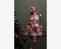 En væghængt dekoration formet som et juletræ, bestående af 24 små stoflommer i røde, hvide og ternede mønstre, hver nummereret fra 1 til 24. Nogle lommer har små gaver eller godter, der stikker ud. Den øverste lomme er pyntet med en sløjfe. Baggrunden er en ensfarvet mørk væg, og til venstre i billedet ses en del af et pyntet juletræ.