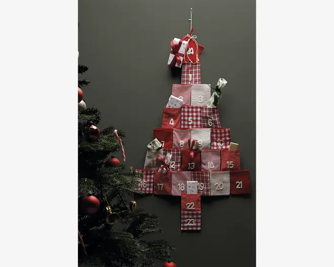 En væghængt dekoration formet som et juletræ, bestående af 24 små stoflommer i røde, hvide og ternede mønstre, hver nummereret fra 1 til 24. Nogle lommer har små gaver eller godter, der stikker ud. Den øverste lomme er pyntet med en sløjfe. Baggrunden er en ensfarvet mørk væg, og til venstre i billedet ses en del af et pyntet juletræ.