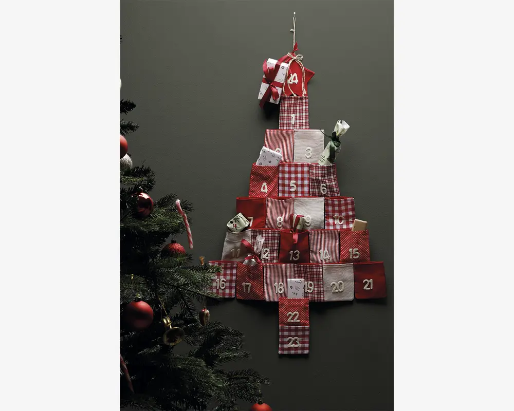 En væghængt dekoration formet som et juletræ, bestående af 24 små stoflommer i røde, hvide og ternede mønstre, hver nummereret fra 1 til 24. Nogle lommer har små gaver eller godter, der stikker ud. Den øverste lomme er pyntet med en sløjfe. Baggrunden er en ensfarvet mørk væg, og til venstre i billedet ses en del af et pyntet juletræ.