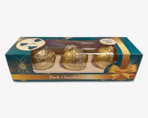 En rektangulær æske med et klart plastikvindue viser tre kugleformede godbidder, hver indpakket i skinnende guldfolie. Æsken er dekoreret i turkis og guld med blomstermønstre og en grafisk guldbånd. Emballagen er elegant og festlig, velegnet som gave.