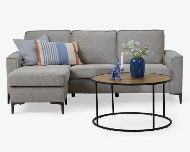 Et moderne stuearrangement med en lysegrå L-formet sofa med tre sædehynder og tre ryghynder. Sofaen har slanke, mørke ben og er pyntet med to pyntepuder: én i ensfarvet lyseblå og én med blå og orange striber. Foran sofaen står et rundt sofabord med en lys træbordplade og en tynd sort metalramme. På bordet står en blå vase med hvide og grønne kunstige blomster, en blå dekorativ dyrefigur, der holder et hvidt stearinlys, samt et tændt lys.