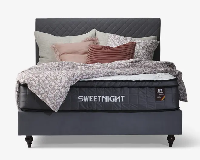 En seng med et mørkegråt, quiltet sengegærde og en tyk madras med mærket 'SWEETNIGHT'. Sengen er redt med et lyse lilla blomstret dynebetræk, flere pyntepuder i beige, rosa og bordeaux nuancer samt et hvidt lagen, der delvist kan ses. Sengerammen er betrukket med matchende mørkegråt stof og står på korte, mørke træben.