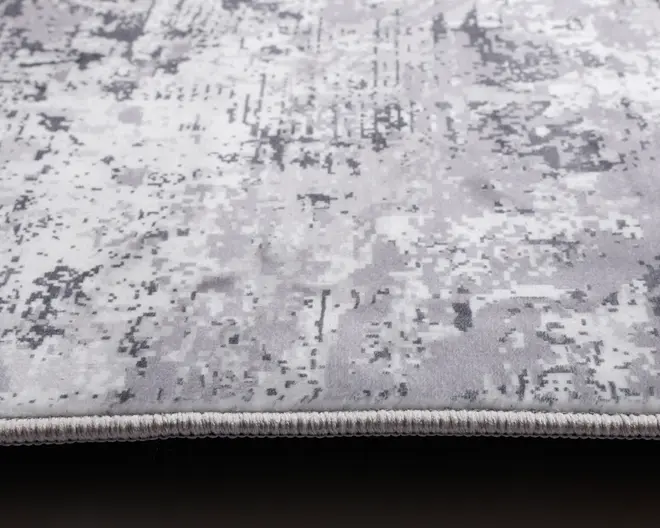 Nærbillede af et tæppe med et moderne abstrakt mønster i grå og hvide nuancer. Overfladen har en blød, fløjlsagtig tekstur med et pixelagtigt design, og kanten er pænt afsluttet med en tæt vævet kant for ekstra holdbarhed.