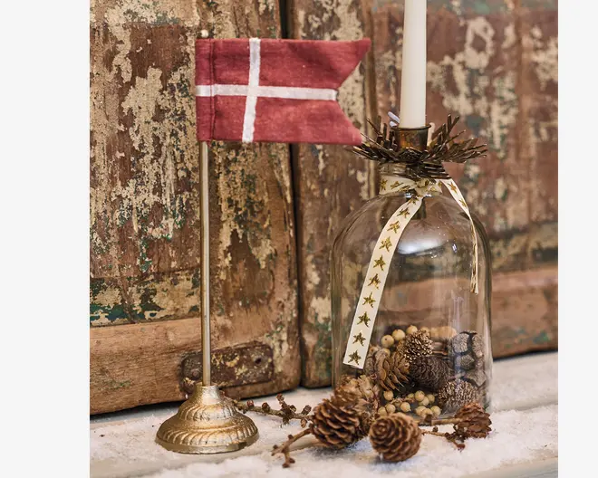 Et lille dekorativt dansk flag på en gylden fod står ved siden af en klar glasklokke med kogler, bær og naturdetaljer, toppet med et hvidt stearinlys og pyntet med et bånd med stjernemønster.